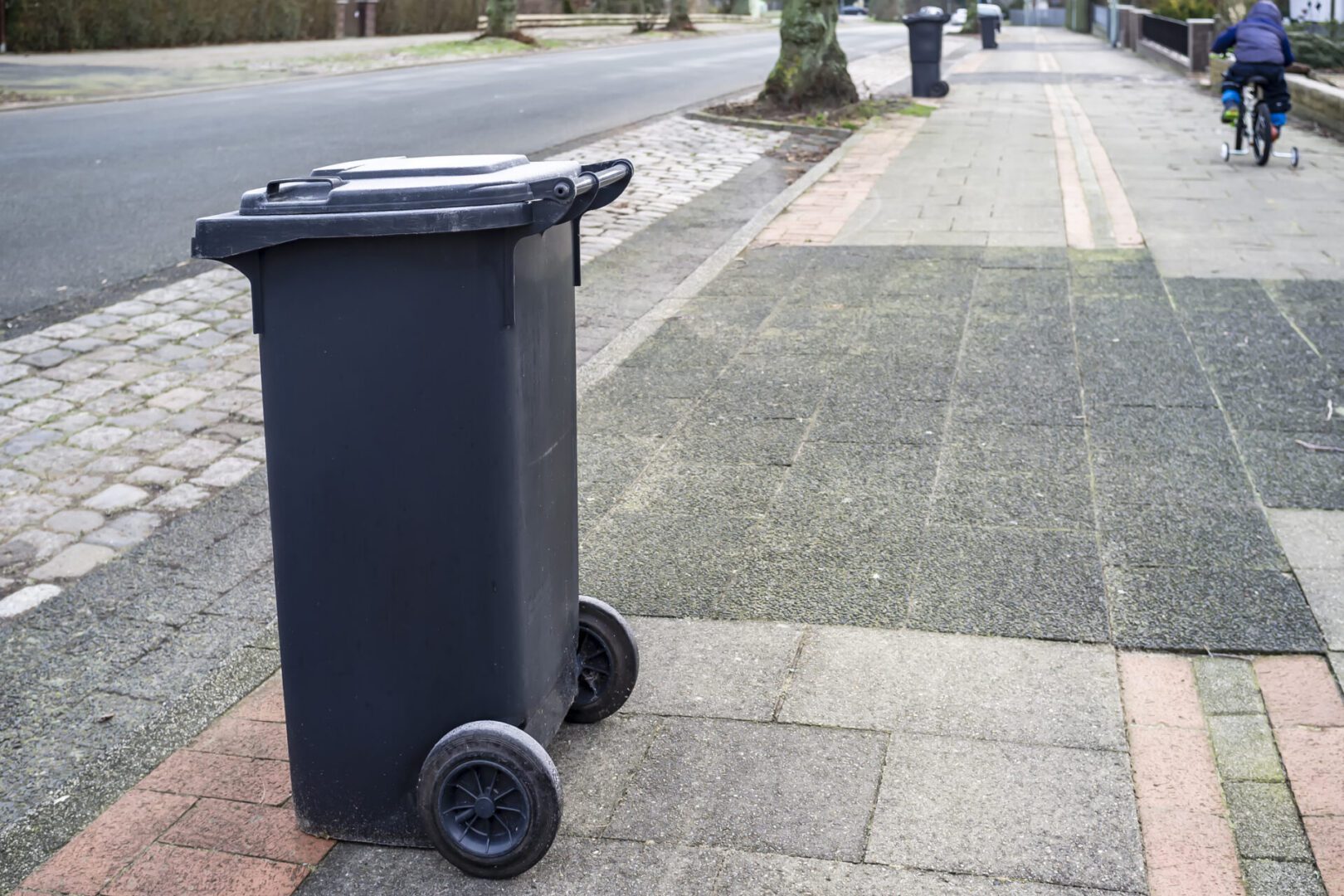 A black outdoor trash bin on wheels near a sidewalk.