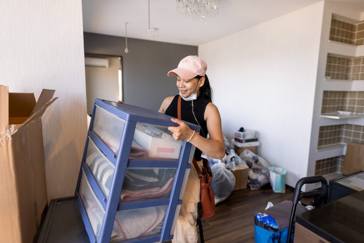 Person organizing items into a blue storage bin in a cluttered room.