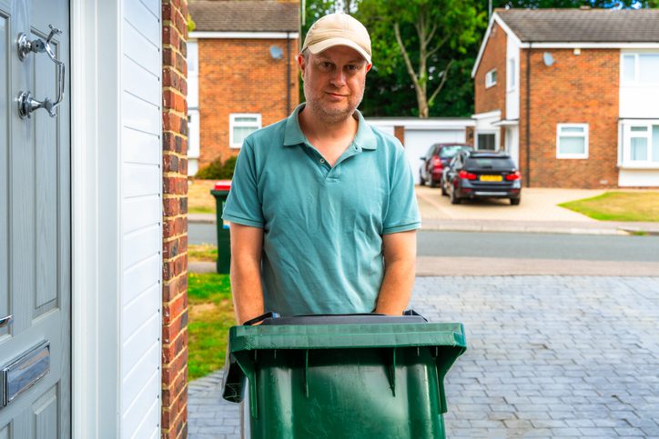 Man standing behind a green trash bin on a residential street.